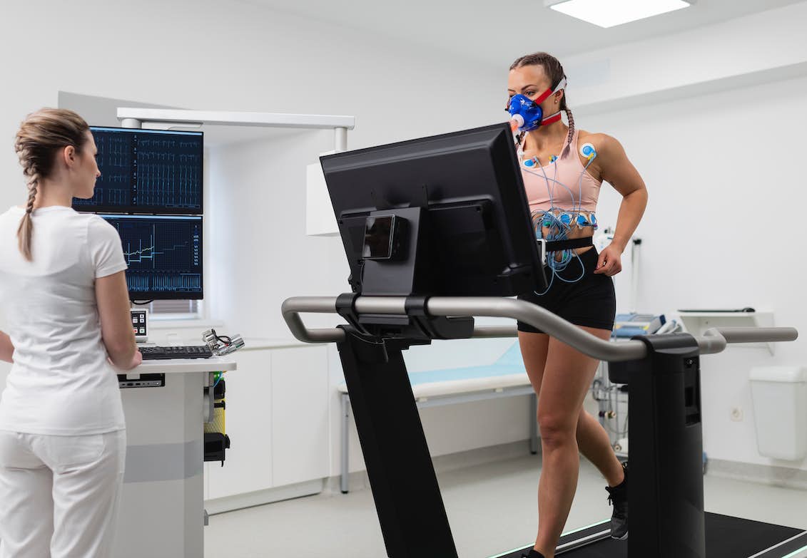 Picture ilustrating how the VO2 max test is perform. There is a lady running in a treadmill, the lady is connected to a machine to measure her levels of oxygen